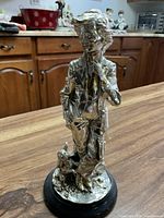 Silver figurine of a boy standing pensively with a dog sitting at his feet on a round black base, shown from an angle highlighting the front and right side.