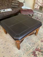 Full view of square coffee table with dark brown vinyl cushioned top and part of the seating area in background showing context.