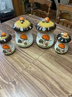 Photo of four mushroom-design ceramic canisters in varying sizes on wood table, showing hand-painted orange and brown mushrooms with green leaves and small orange ladybug accents on the lids.