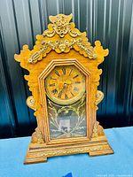 Front view of antique gingerbread mantle clock showing full wooden case with carved gold-tone trim, original glass door, and clock face with floral design and Roman numerals.