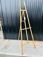 Full view of the folding pine wood artists easel standing upright outdoors against a dark corrugated metal background, showing the overall structure and finish.