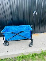Front and side view of blue fabric wagon with black metal frame, four wheels, and extended handle on concrete ground with black wall background.