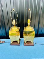 Pair of brass Chinese table lamps displayed side by side, showing square brass bodies on rosewood bases, with lamp fittings at top.