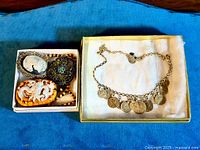 Box with three brooches including two cameos and one filigree with stones, and adjacent gold-tone necklace with coin pendants.