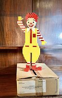 Front view of LEGO Ronald McDonald figure standing on shipping box on wooden shelf