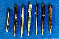 Overhead photo showing seven vintage pens and pencils arranged side by side on a blue cloth, showcasing the different finishes and colors.