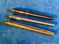 Three Sheaffer fountain pens: gold-plated, silver-tone, and pearlescent dark blue, laid on blue fabric.