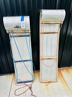 Photo showing two aluminum vintage toboggans side by side, one with blue rope and the other with red and white rope attached, each having wood slats for seating.