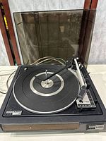 Front angled view showing black turntable with circular silver platter, tonearm, and dark-tinted dust cover, with cords visible in background.