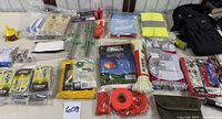 Full assortment of camping and survival gear spread on a table showing bag, ropes, stakes, rain ponchos, safety vest, whistle, thermal blankets, tent, and various small items
