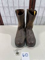 Front view of worn brown leather Caterpillar work boots with CAT logo tag in bag and lot number card.