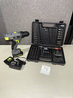 Master Mechanic 12V lithium cordless drill with battery and charger alongside Black & Decker drill bit set in black case on table.
