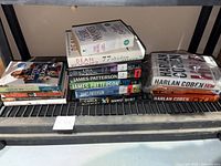 Shelf with multiple used books by authors Harlan Coben, James Patterson, Dean Koontz, and others, stacked horizontally and vertically.