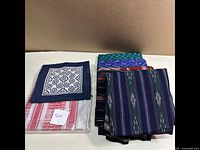 Photo showing folded fabrics including a prominent navy square with white swirl design, striped and ikat fabrics in blue, green, and red tones folded and stacked.