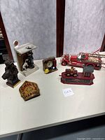 Various firefighter collectible items: vintage red fire truck phone, firefighter bust figurine with Dalmatian, fire station coin bank, firefighter patch, small firefighter figurine, fire hydrant clock, and badge plaque.