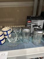 OXO SoftWorks French Press in box, two glass sugar containers with metal lids, two clear glass coffee cups with glass saucers, and two ceramic mugs with blue and beige checkered pattern