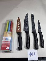 Photo showing three Millerhaus black speckled non-stick kitchen knives next to a copper-colored kitchen knife with black handle and the boxed 7-piece Klok cutlery steak knife set with multicolored handles.