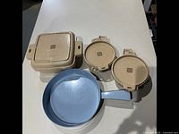 Three beige ceramic cookware pieces with lids and an 8-inch light blue skillet displayed on white table.