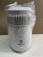 Front view of white Megahome water distiller wrapped in plastic, showing ventilation slats and logo.