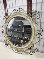 Front view of round metal framed mirror showing floral and scroll metal decorations with distressed white and yellow paint finish.