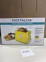 Box for Nostalgia Grilled Cheese Sandwich Toaster showing toaster image, yellow color, and features such as adjustable toasting dial, making two sandwiches at once without mess.