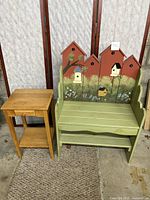 Wooden bench painted green with birdhouse and nature scene on backrest, next to natural wood small table with drawer and lower shelf, placed on carpet.