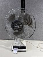 Front view of Panasonic oscillating fan showing metal grille and blades, white base with control panel.