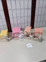 Four Precious Moments figurines posed in front of their original boxes in yellow, peach, and pink colors. Each figurine shows soft pastel colors and detailed companions or objects.