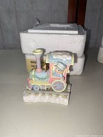 Photo of pastel colored Precious Moments train engine figurine with conductor bear on a track base, beside original white Styrofoam packaging.