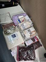 Lot of queen size sheets and valances in various colors arranged on table, mostly packaged and new, including purple duvet set and 600 thread count sheets.