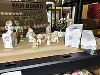 Photo showing an assortment of small figurines including babies, girls, and a heart-shaped photo holder arranged on a cardboard surface with some items in boxes.