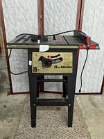 Front view showing the table saw standing on black metal legs, control panel with labels and adjustment knobs visible.