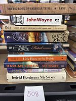 Stack of assorted used paperback and hardcover books including titles by John Wayne, Stephen King, Marie Force, and Ingrid Bergman