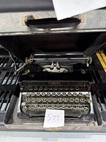 Black Corona Junior typewriter with silver-edged round keys and white lettering inside a black carrying case with fabric lining.