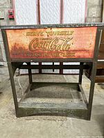 Front view showing rusted and faded Coca Cola advertising with 'Serve Yourself, Drink Coca-Cola Please Pay the Clerk' text on metal panel of cooler.