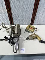 All four vintage power tools placed on white surface showing overall lot contents with power cords visible.