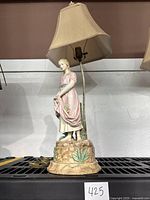 Full view of the porcelain figural lamp showing the female statue base standing on a decorative round base with painted leaves and fabric lampshade.