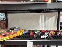 Wide view photo showing vintage games and assorted toy vehicles including a Tic Tac Dough game box, various toy vehicles such as fire trucks, cars, and helicopters arranged on a shelf.