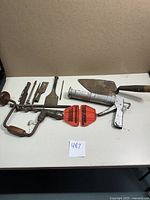 All items displayed on table showing antique hand drill, wooden handle trowel, metal chisels, red plastic drill bit holder, and the halogen leak detector case assigned in background.