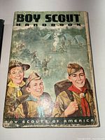 Front cover of the vintage Boy Scout Handbook with an illustrated cover depicting three Boy Scouts and camp scenes, showing wear and tear.