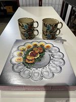 Four vintage stoneware mugs with floral design stacked in pairs next to a boxed crystal egg relish server on a white table