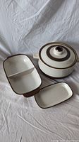 Three Denby cookware pieces pictured on white fabric: a lidded pot and two rectangular dishes, one divided.