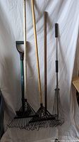 Four yard tools including three leaf rakes and one garden cultivator with green handle and D-grip against a white backdrop.