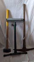 Full view of the sledgehammer, pickaxe, and manual construction roller standing upright on a white cloth background.