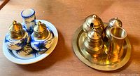 Blue and white ceramic salt, pepper, and toothpick set on a matching ceramic tray next to a brass salt, pepper, toothpick, and coarse salt set on a brass round tray on a wood table