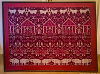Framed tapestry showing full fabric with white animal, human, geometric, and architectural motifs on red background.