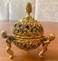 Front view of the closed gold tone ornate jewelry holder with cherub supports and pinecone finial on lid.