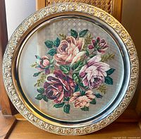 Front view showing round decorative glass serving tray with gold ornate rim and mounted needlepoint floral picture inside glass dome.
