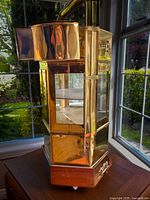 Front angle of tall brass noodle cart with glass windows and curved top spout, sitting on wooden wheeled base.