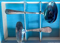 Two spreading utensils placed in a box, one with a ladle-like rounded spoon head and the other with a flat, wide blade, both metal with tarnishing visible.